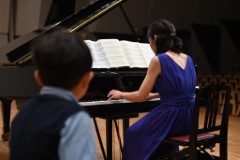 ピアノの斜め後ろの椅子に座った子どもを前景に、その視線の先でピアノを弾く母の後ろ姿を写した一枚。